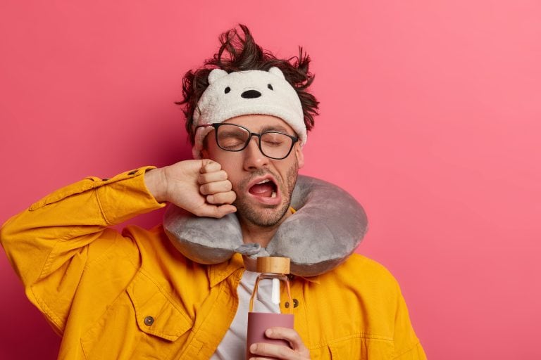 photo of exhausted unshaven man traveler yawns from tiredness, wants sleep badly, holds glass bottle of drink, wears neck pillow, awakes during long trip in plane, isolated over pink background